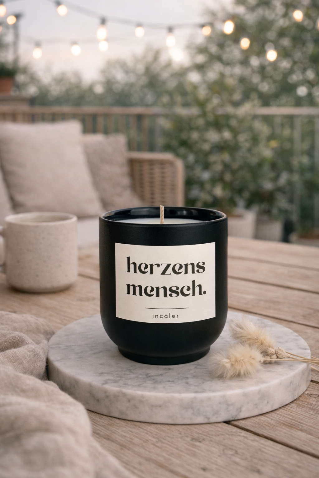 Schwarze (basalt) Kerze mit der Aufschrift ‚herzensmensch‘ steht auf einer runden Marmorplatte auf einem Holztisch auf dem Balkon. Im Hintergrund sind ein Sitzbereich, Pflanzen und eine Lichterkette zu sehen, die eine warme, entspannte Atmosphäre schaffen.
