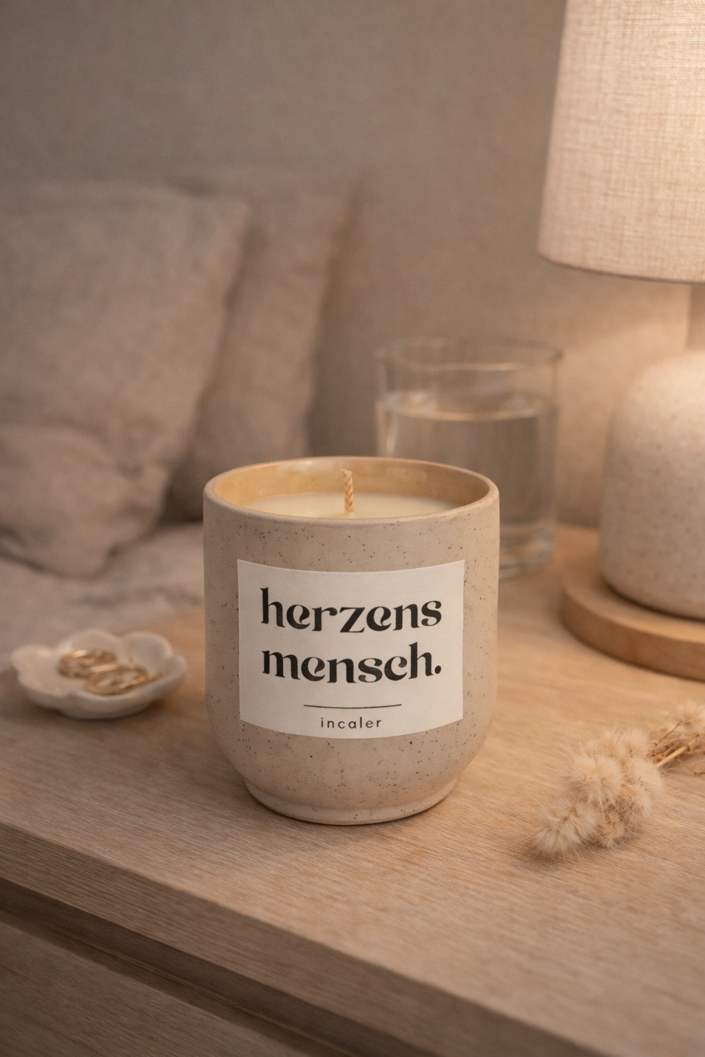 Beige (sahara), gesprenkelte Kerze mit der Aufschrift ‚herzensmensch‘ steht auf einem Holztisch neben einer Lampe, einem Glas Wasser und kleinen Dekoelementen. Warmes Licht schafft eine gemütliche, ruhige Atmosphäre.