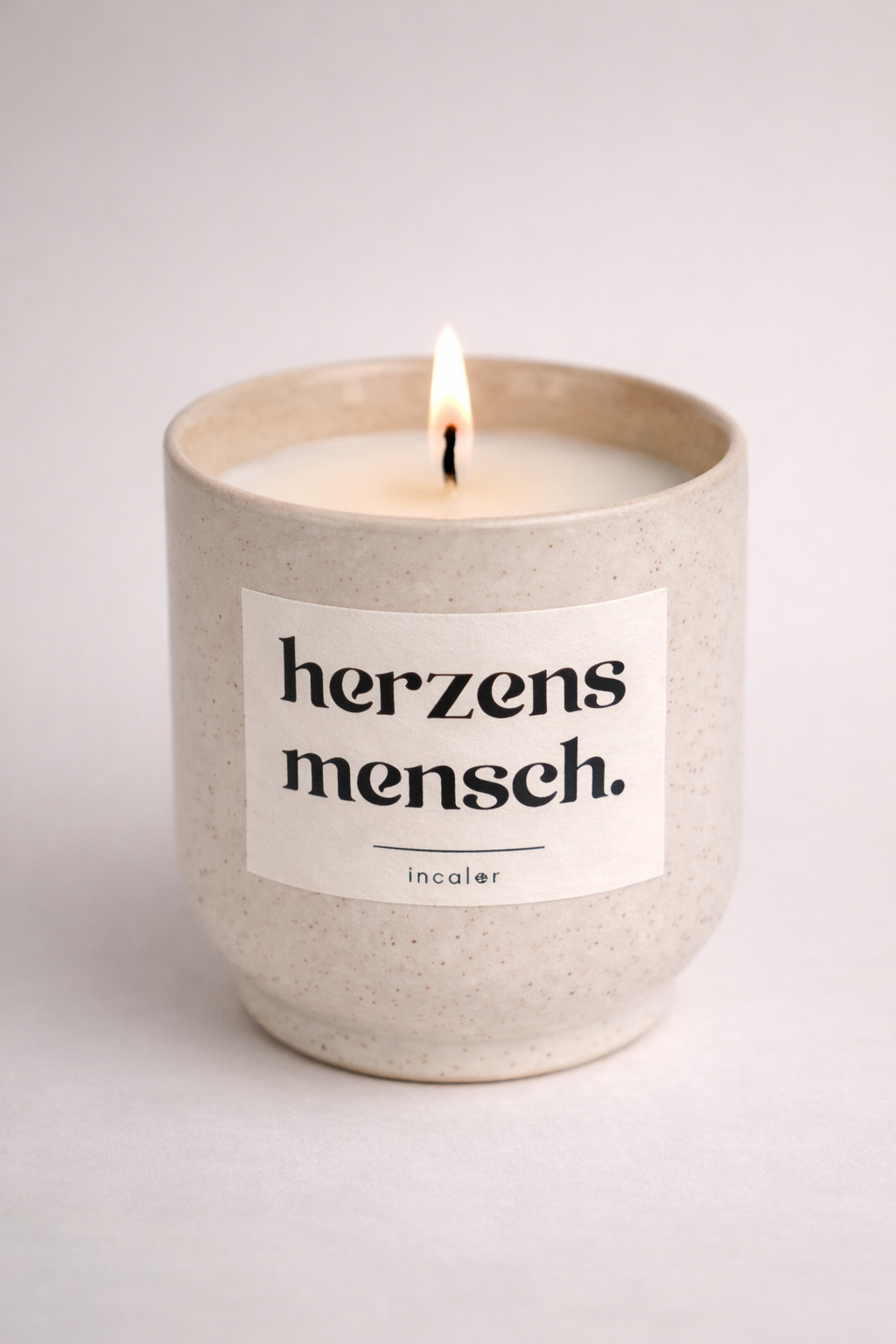 Helle, beige (sahara) gesprenkelte Kerze mit der Aufschrift ‚herzensmensch‘ brennt vor einem neutralen Hintergrund. Die Flamme ist sichtbar und sorgt für eine ruhige, minimalistische Atmosphäre.