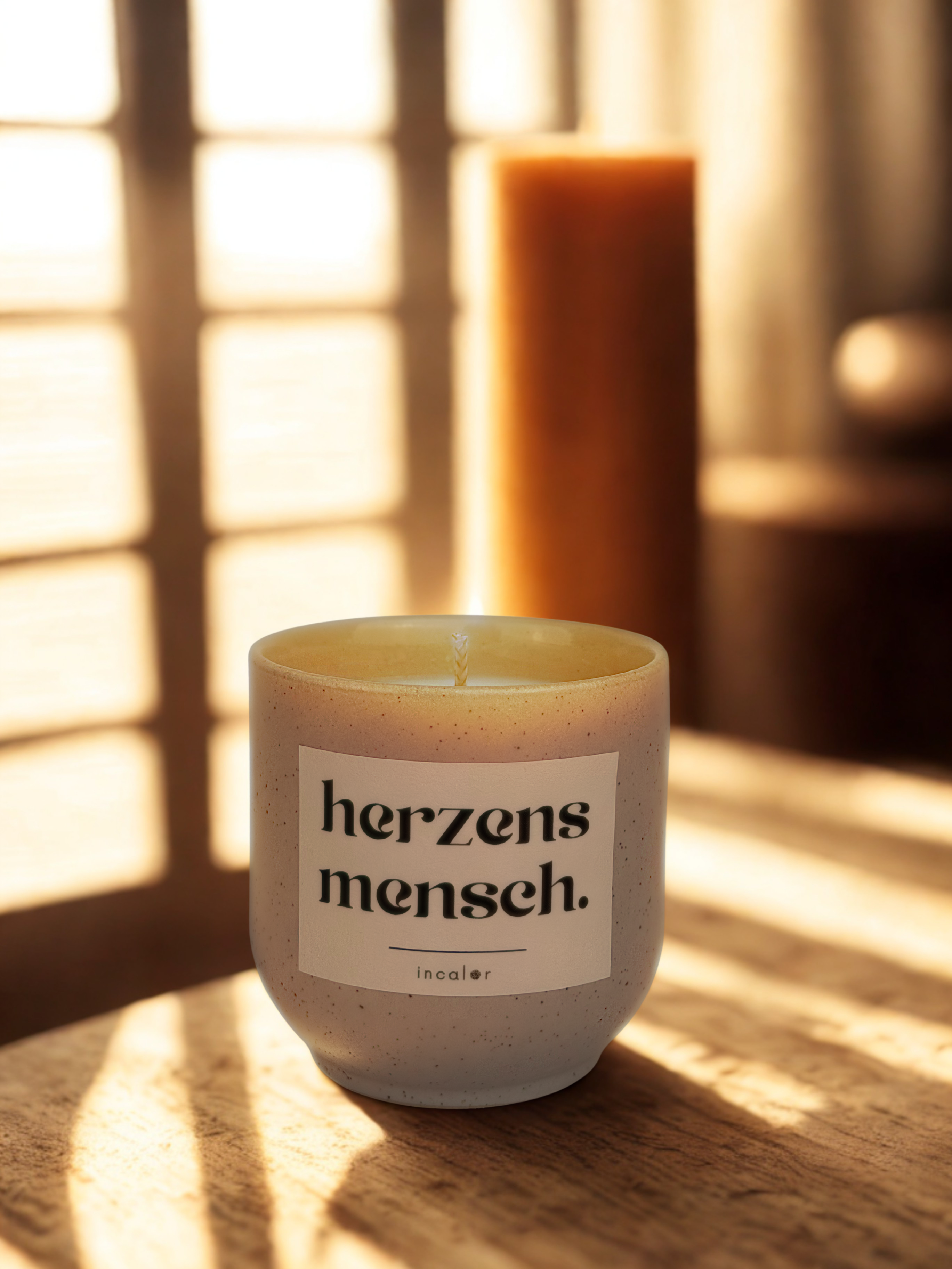 Helle, cremefarbene (sahara) Keramikduftkerze mit dem Schriftzug „herzensmensch.“ im minimalistischen incalor-Design, stimmungsvoll arrangiert im warmen Sonnenlicht eines gemütlichen Raumes.