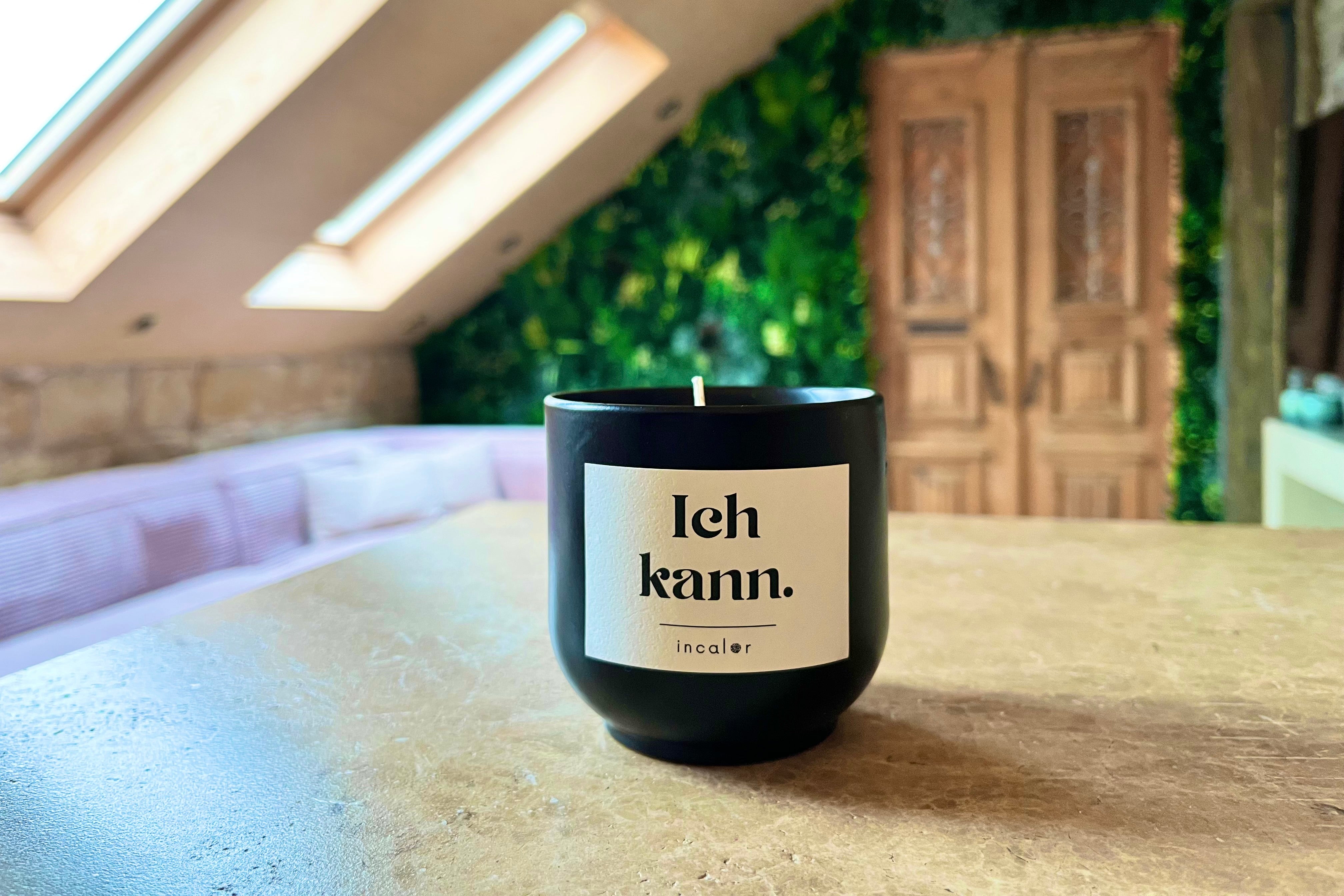 Eine schwarze Duftkerze mit weißem Etikett steht auf einem hellen Tisch. Auf dem Etikett steht: „Ich kann. – incalor“. Im Hintergrund ist ein gemütlicher Raum mit Dachschrägen, großen Dachfenstern, einer grünen Pflanzenwand und einer rustikalen Holztür zu sehen. Das Licht fällt warm durch die Fenster.