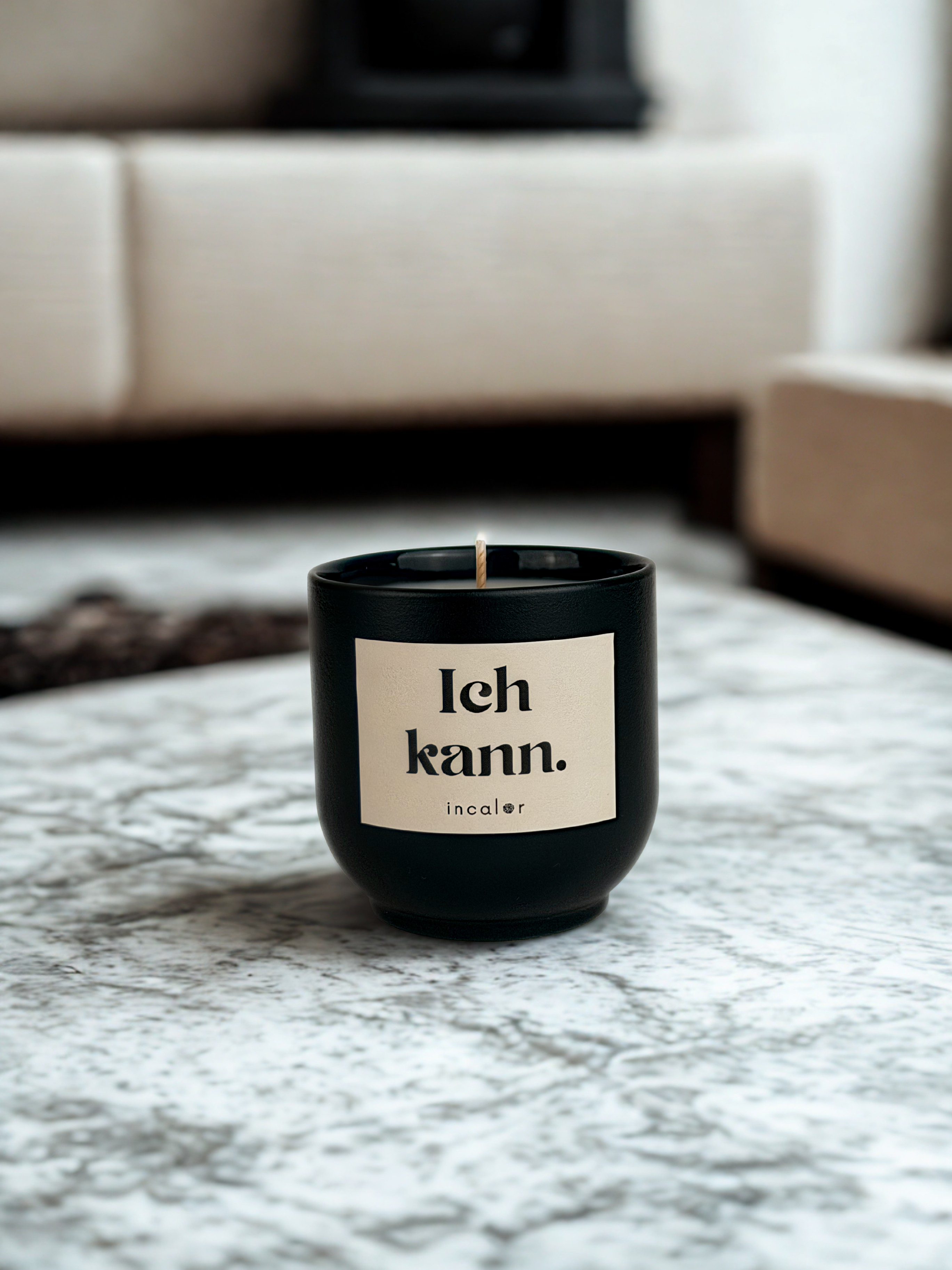 Eine Kerze in der Farbe grün (deep ivy) und Etikett mit der Aufschrift "Ich kann" steht schön platziert auf einem Steintisch in einem Wohnzimmer.