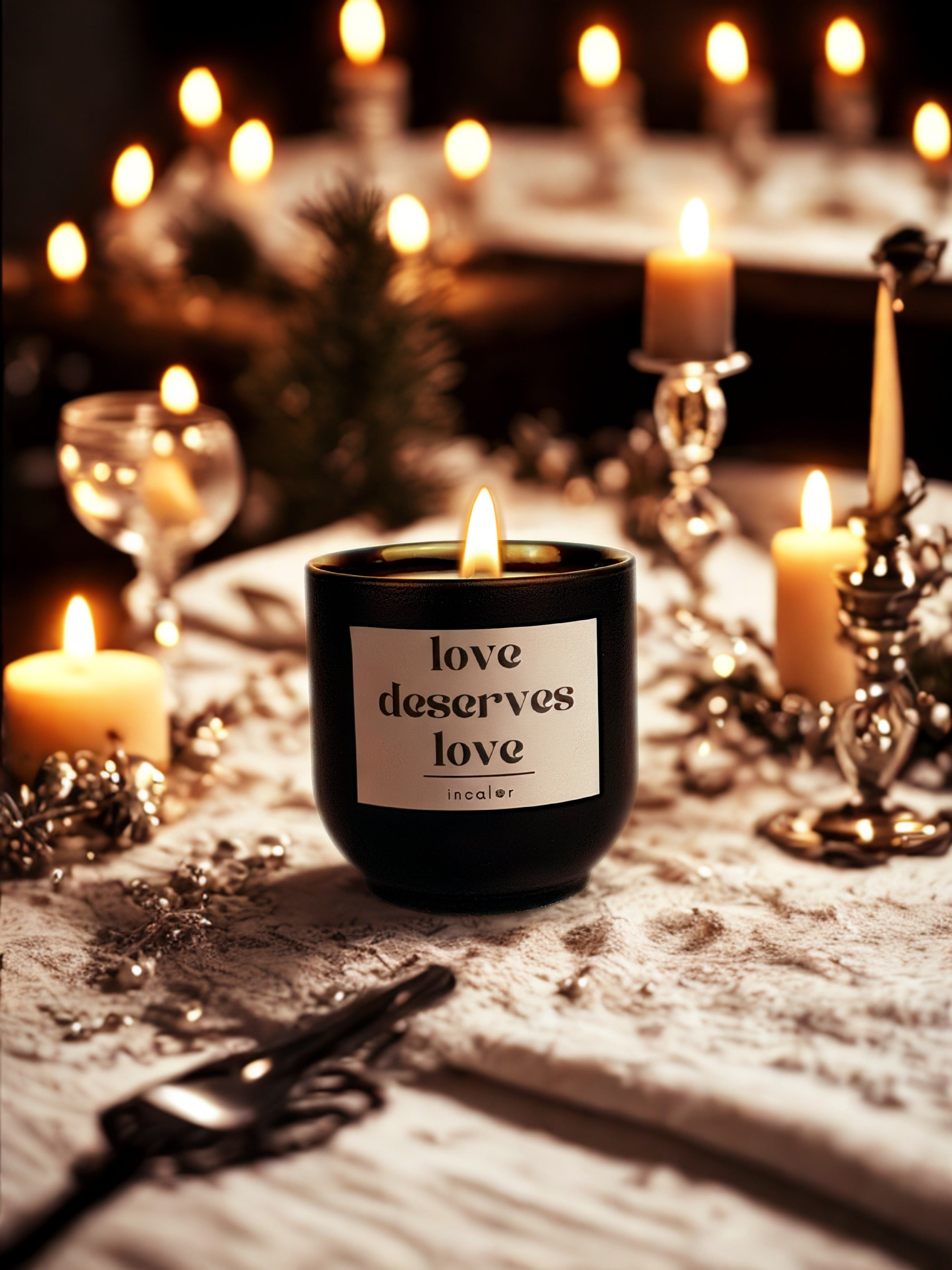 Schwarze Duftkerze mit brennender Flamme steht auf einem festlich dekorierten Tisch. Auf dem Etikett steht „love deserves love“ mit dem Markennamen „incaler“. Umgeben ist die Kerze von mehreren warm leuchtenden Stumpenkerzen, silberner Tischdekoration und einem edlen Besteck. Im Hintergrund sorgen weitere Kerzen und weiche Lichter für eine romantische, elegante Atmosphäre.