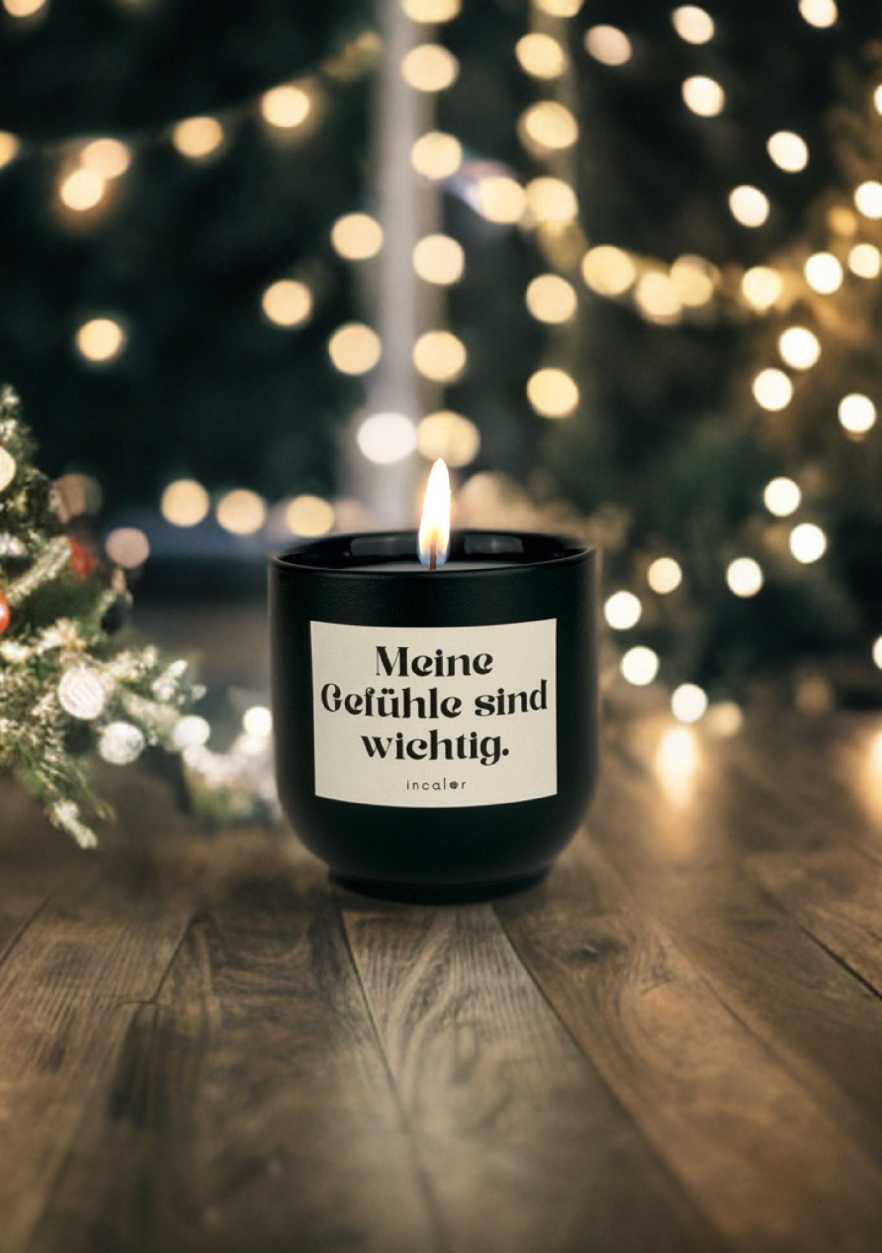 Dunkelgrüne (deep ivy) Duftkerze mit brennender Flamme steht auf einem dunklen Holzboden. Auf dem Etikett steht „Meine Gefühle sind wichtig.“ mit dem Markennamen „incaler“. Im Hintergrund leuchten unscharfe, warme Lichter eines Weihnachtsbaums und Tannenzweige sorgen für eine festliche Atmosphäre.