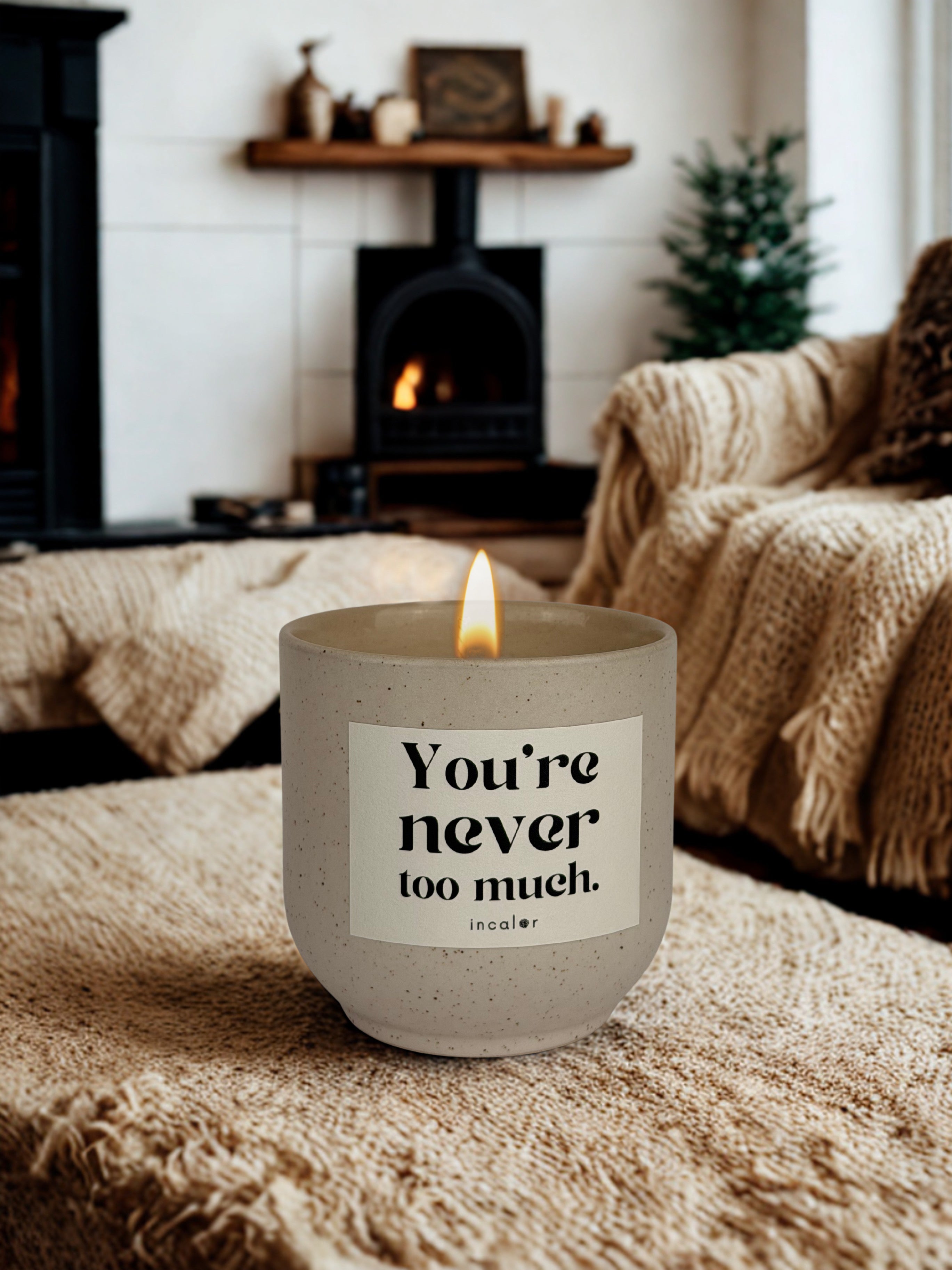 Beige (sahara) Duftkerze mit brennender Flamme steht auf einer weichen, grob gestrickten Decke in einem gemütlichen Wohnzimmer. Auf dem Etikett steht „You're never too much.“ mit dem Markennamen „incalor“. Im Hintergrund befinden sich ein Kamin mit Feuer, ein Sofa mit beigefarbenen Decken und ein kleiner Weihnachtsbaum, die eine warme, heimelige Atmosphäre schaffen.