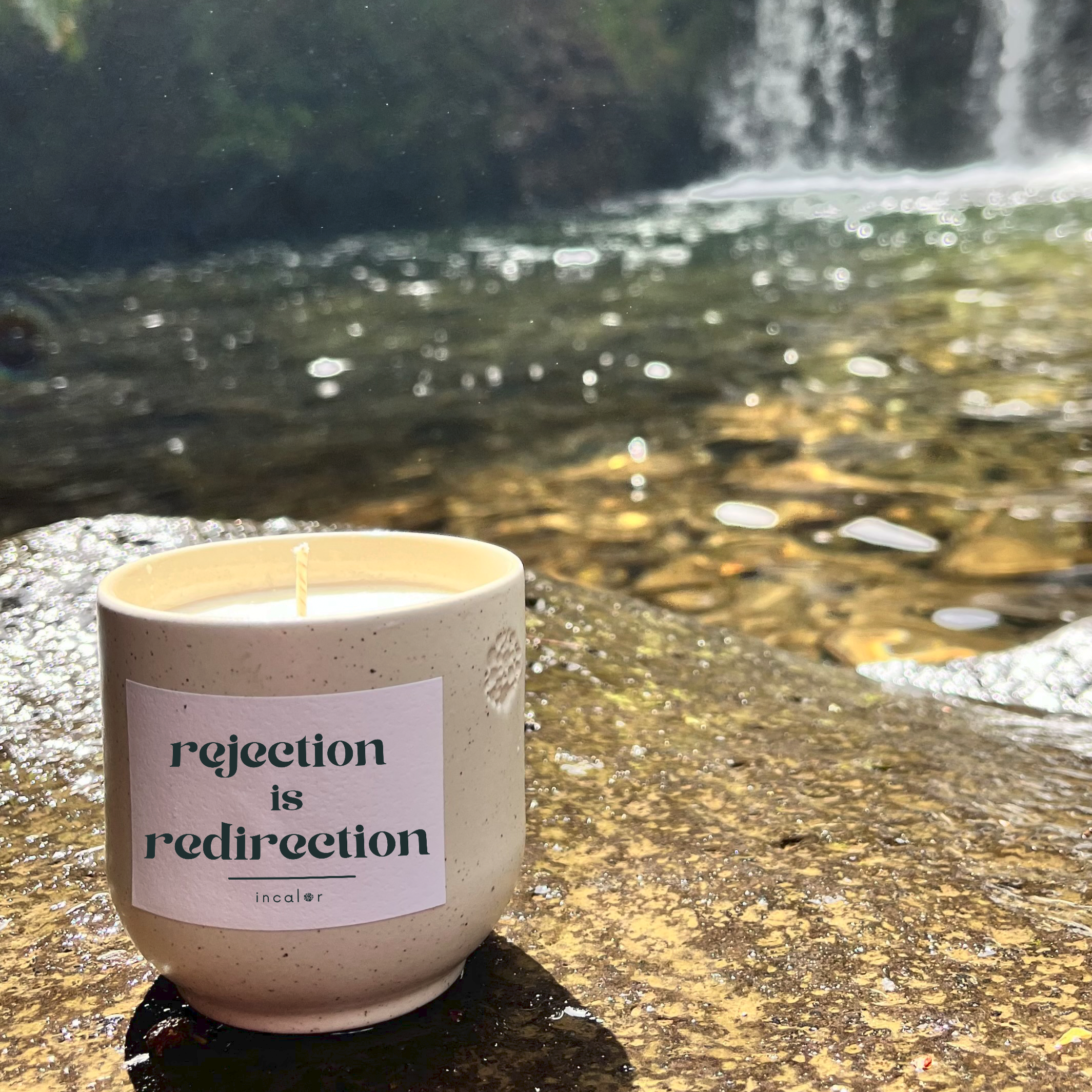 Kerze aus beige Keramik (Farbton sahara) mit Label der Affirmation "rejection is redirection", stilvoll platziert auf Naturstein mit einem Wasserfall im Hintergrund – hochwertige Designkerze für modernes, minimalistisches Wohndesign.