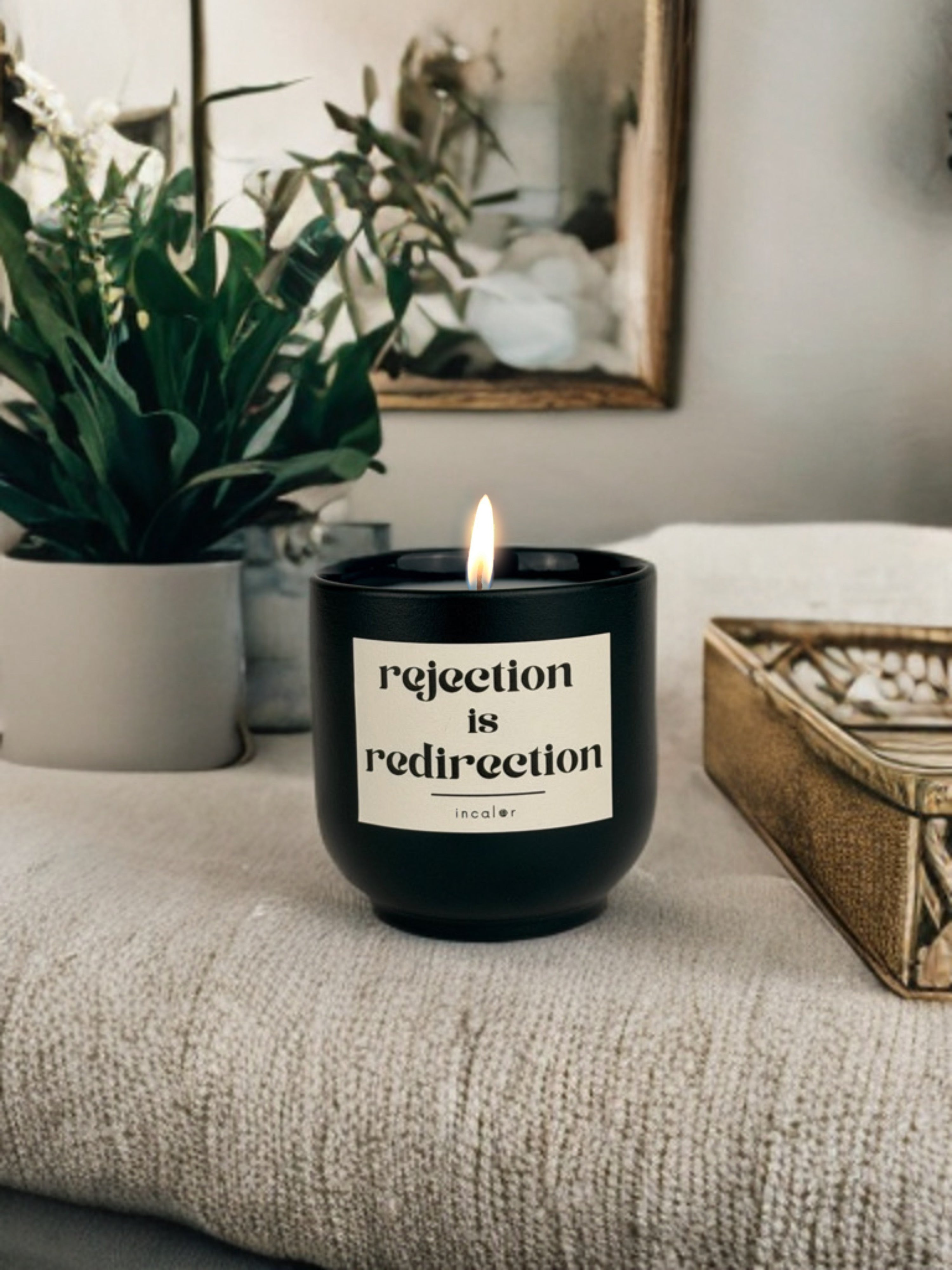 Dunkelgrüne (deep ivy) Duftkerze mit brennender Flamme steht auf einer beigefarbenen, strukturierten Decke. Auf dem Etikett steht „rejection is redirection“ und darunter der Markenname „incalor“. Im Hintergrund befinden sich eine grüne Zimmerpflanze in einem Topf, ein goldfarbener Aufbewahrungskorb und ein Spiegel, die eine warme, gemütliche Atmosphäre erzeugen.