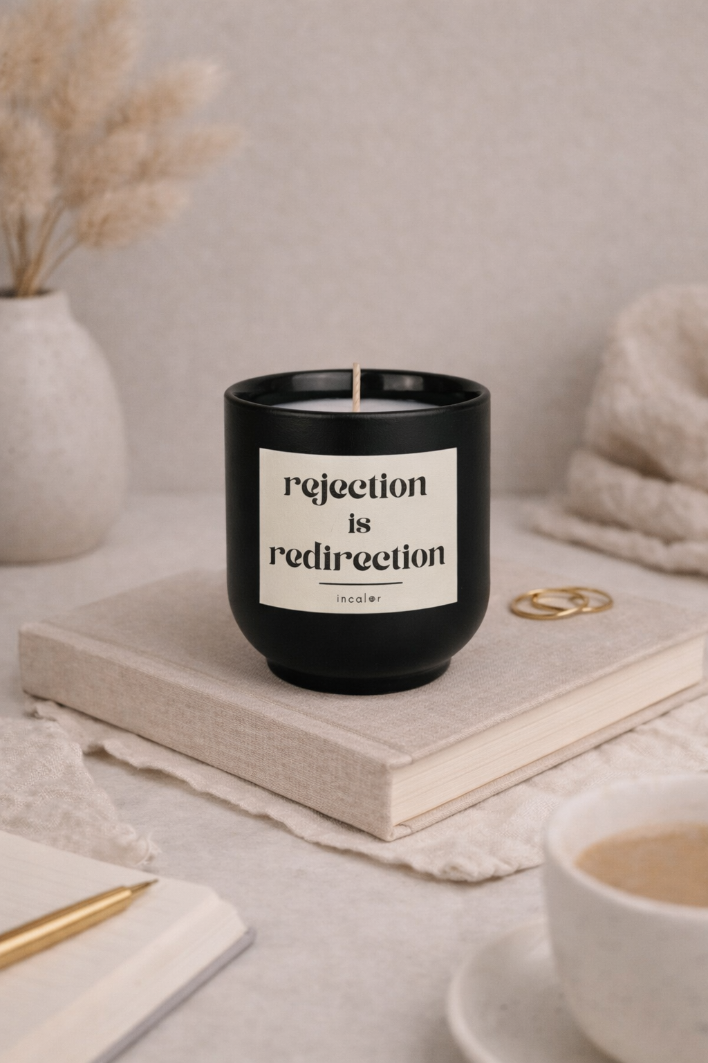 Schwarze (basalt) Duftkerze mit der Aufschrift ‚rejection is redirection‘ steht auf einem geschlossenen Buch. Umgeben von einer Vase mit Trockenblumen, Schmuckringen, einem Notizbuch mit Stift und einer Tasse entsteht eine ruhige, warme und minimalistische Wohnatmosphäre.