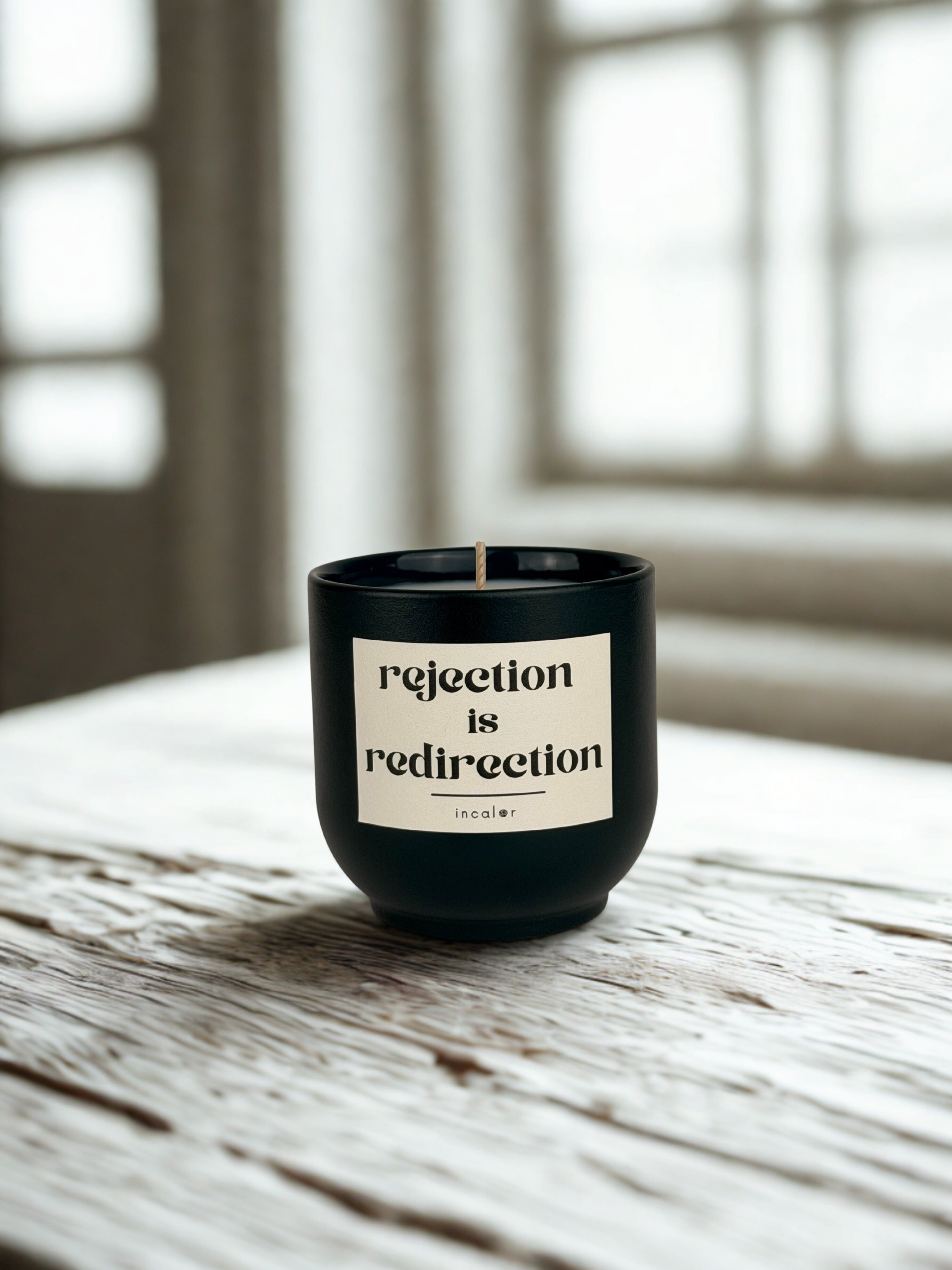 Dunkelgrüne (deep ivy) Duftkerze mit der Aufschrift ‚rejection is redirection‘ steht auf einem rustikalen Holztisch vor einem hellen Fenster. Die Szene wirkt ruhig, minimalistisch und natürlich durch das weiche Tageslicht.