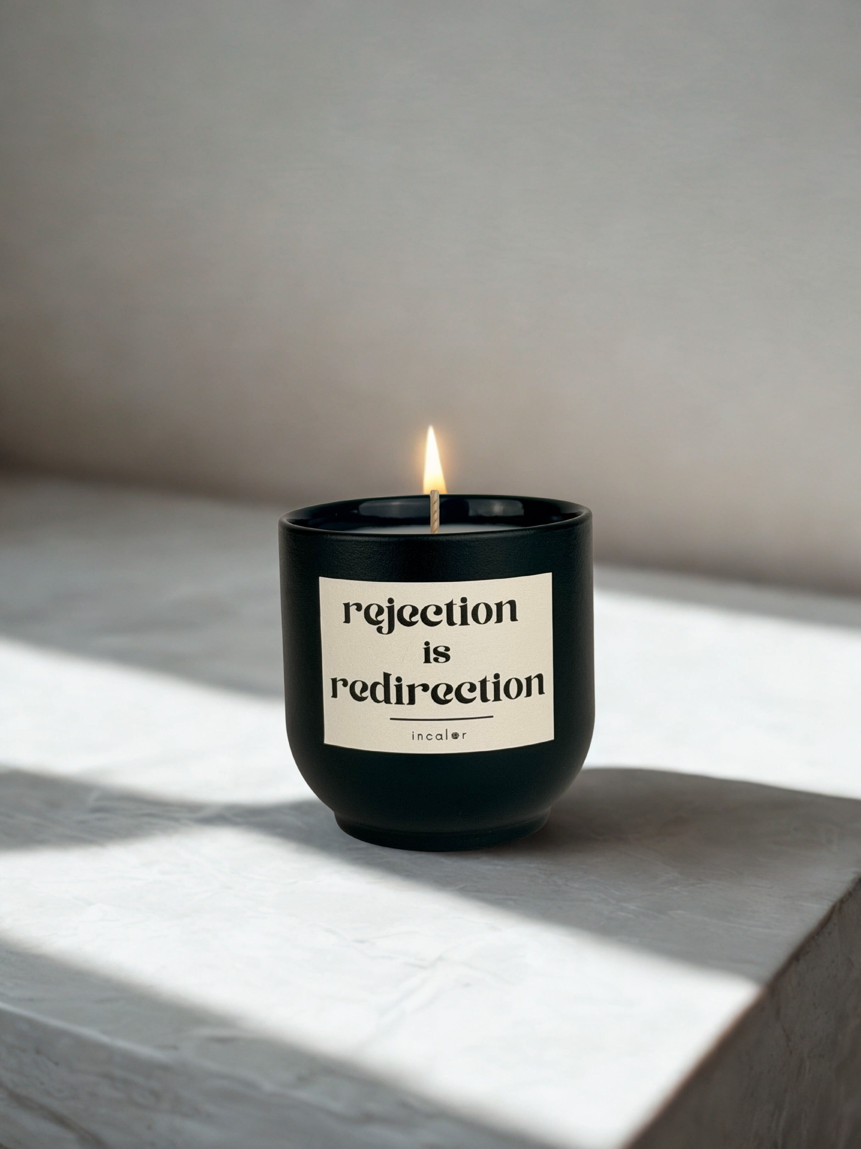 Brennende dunkelgrüne (depo ivy) Duftkerze mit der Aufschrift ‚rejection is redirection‘ steht auf einer hellen Steinoberfläche. Sanftes Sonnenlicht fällt schräg ein und erzeugt eine ruhige, minimalistische und warme Stimmung.
