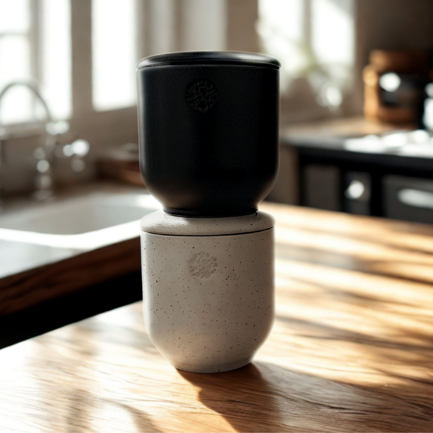 Zwei Keramikgefäße stehen übereinander auf einem sonnendurchfluteten Holztisch in einer Küche. Unten ein beiger (sahara), gesprenkelter Becher mit Deckel, darauf ein schwarzer (basalt) Becher; beide mit einer kleinen runden incalor Logo-Prägung auf der Vorderseite.
