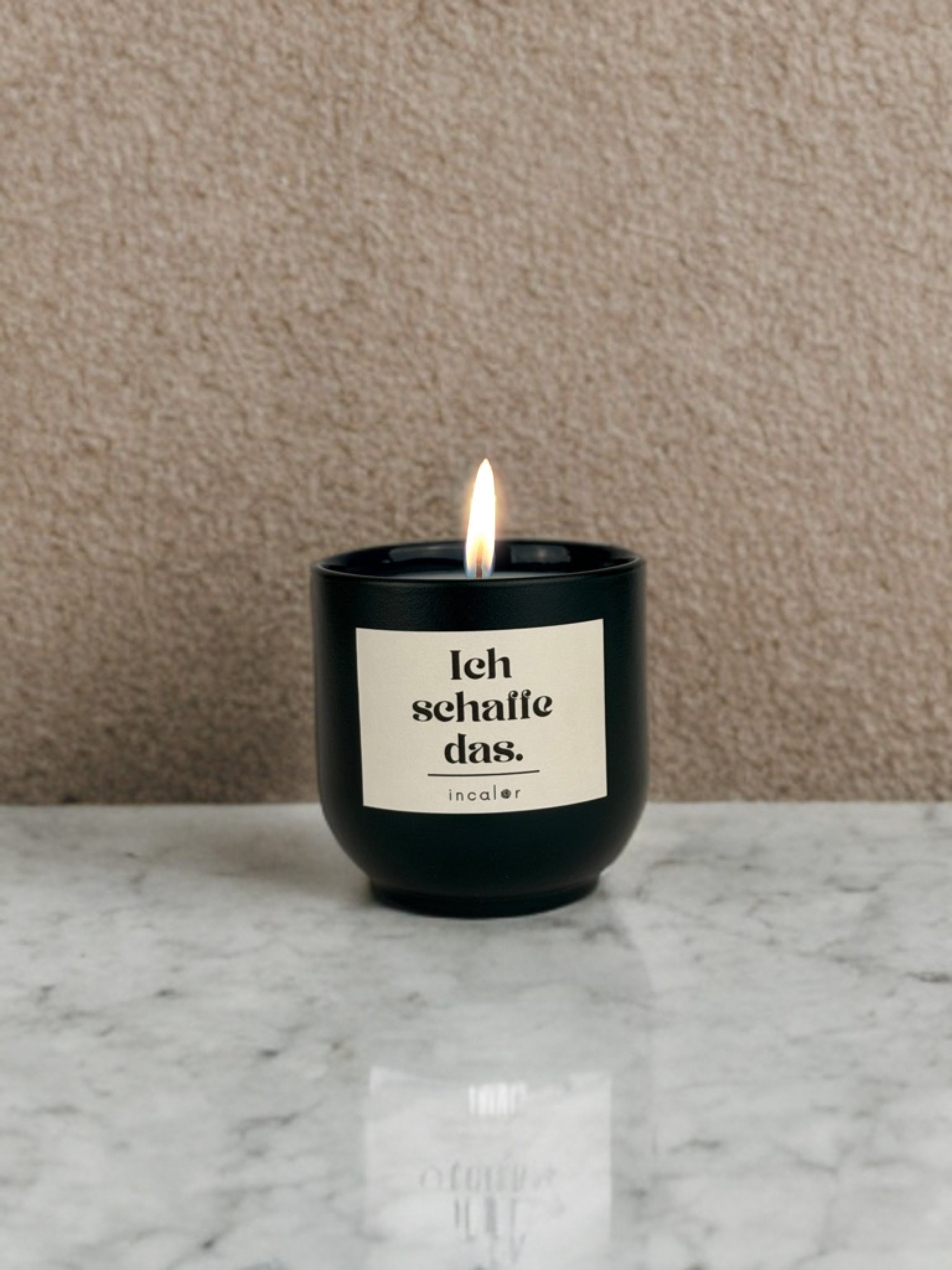 Dunkelgrüne (deep ivy) Duftkerze auf einer hellen Marmoroberfläche, mit brennender Flamme. Auf der Vorderseite steht ein cremefarbenes Etikett mit dem Text „Ich schaffe das.“ und dem Markennamen „incalor“ darunter; Hintergrund ist eine beige strukturierte Wand.