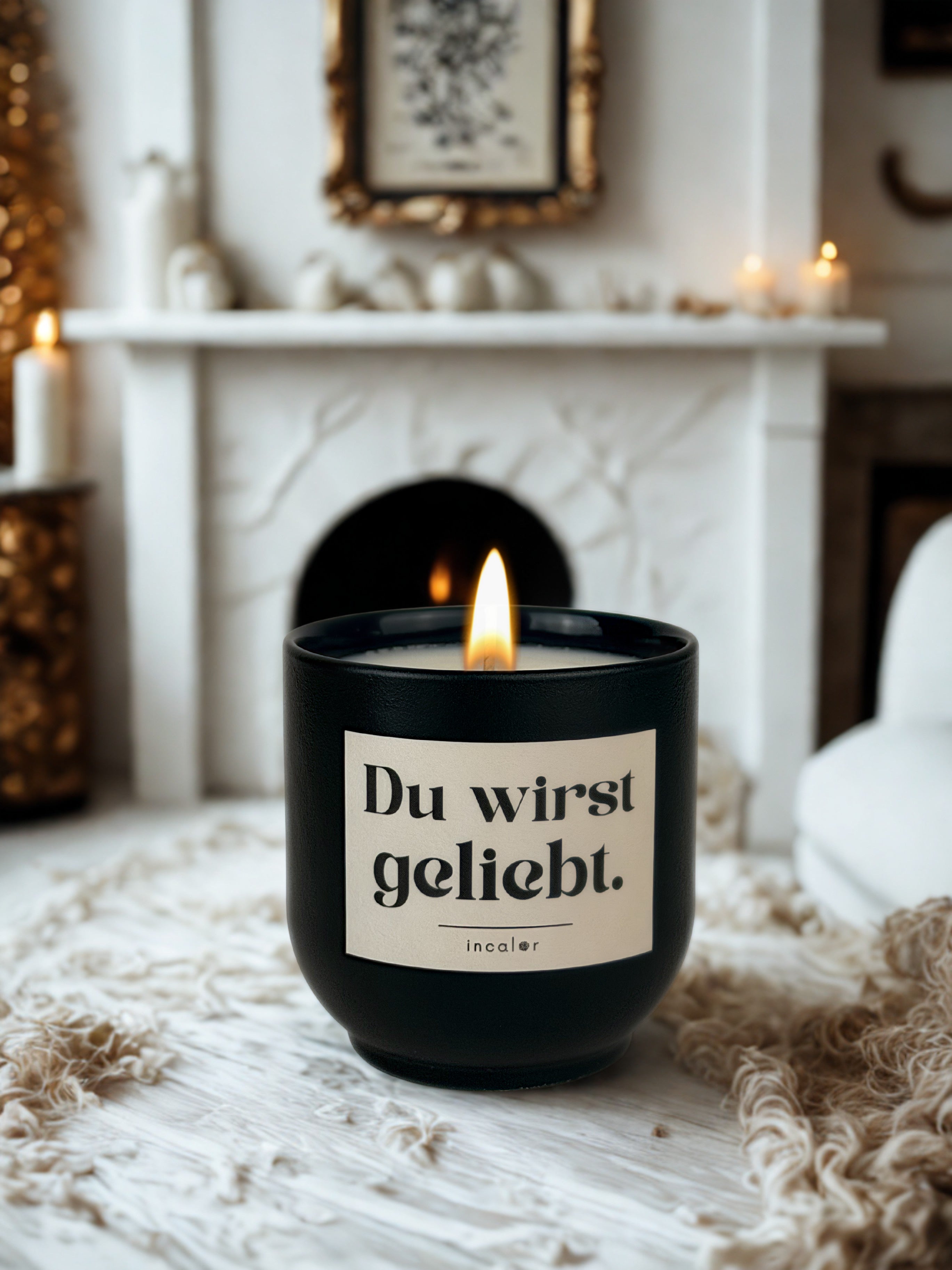 Dunkelgrüne (deep ivy) Duftkerze mit brennender Flamme steht auf einer hellen, weichen Decke in einem gemütlichen Wohnzimmer. Auf dem Etikett steht „Du wirst geliebt.“ und darunter der Markenname „incalor“. Im Hintergrund ist ein Kamin mit weißer Dekoration, Kerzen und einem goldgerahmten Bild zu sehen, wodurch eine warme, behagliche Atmosphäre entsteht.