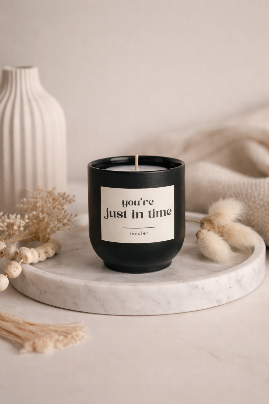 Schwarze (basalt) Kerzen-Tasse mit der Aufschrift ‚you’re just in time‘ steht auf einem runden Marmortablett. Umgeben von Trockenblumen, Holzperlen und weichen Textilien entsteht eine ruhige, natürliche Wohlfühlatmosphäre.