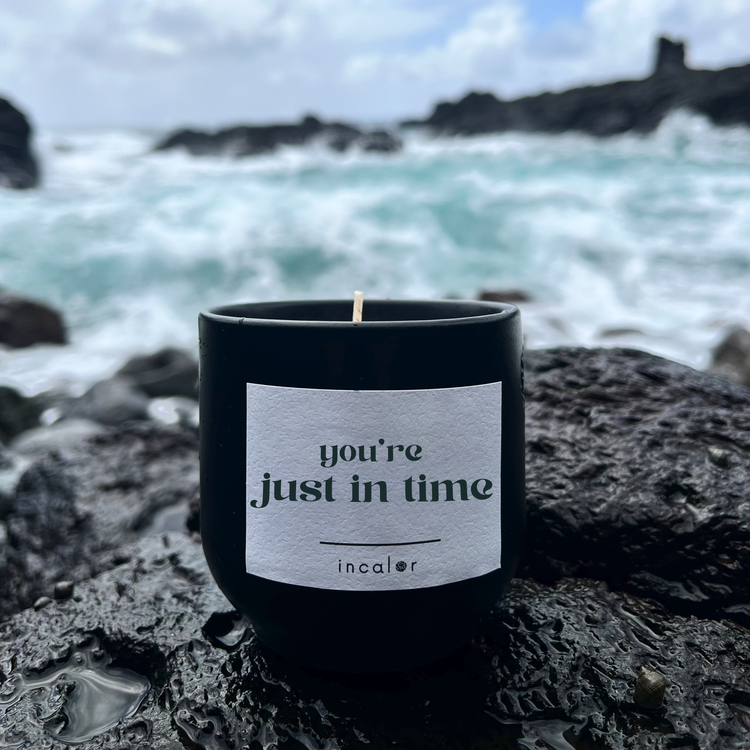 Schwarze Kerze aus Keramik (Farbton Basalt) mit Affirmation „You're just in time.“ von incalor, auf nassem Lavagestein am Meer platziert. Im Hintergrund branden Wellen gegen Felsen unter wolkigem Himmel – Symbol für Resilienz und Achtsamkeit.